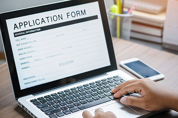 A person types on a laptop with an application form displayed on the screen. A smartphone rests on the table beside the laptop.