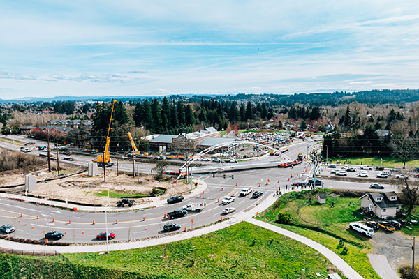 Aerial view of a busy intersection under construction, with vehicles, traffic cones, a crane, and surrounding trees in a suburban area.