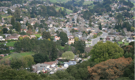 Aerial view of a suburban neighborhood with houses, winding roads, and surrounded by lush green trees and hills.