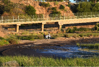 Three people stand on a dry riverbed near a low bridge, surrounded by sparse vegetation and dry grass.