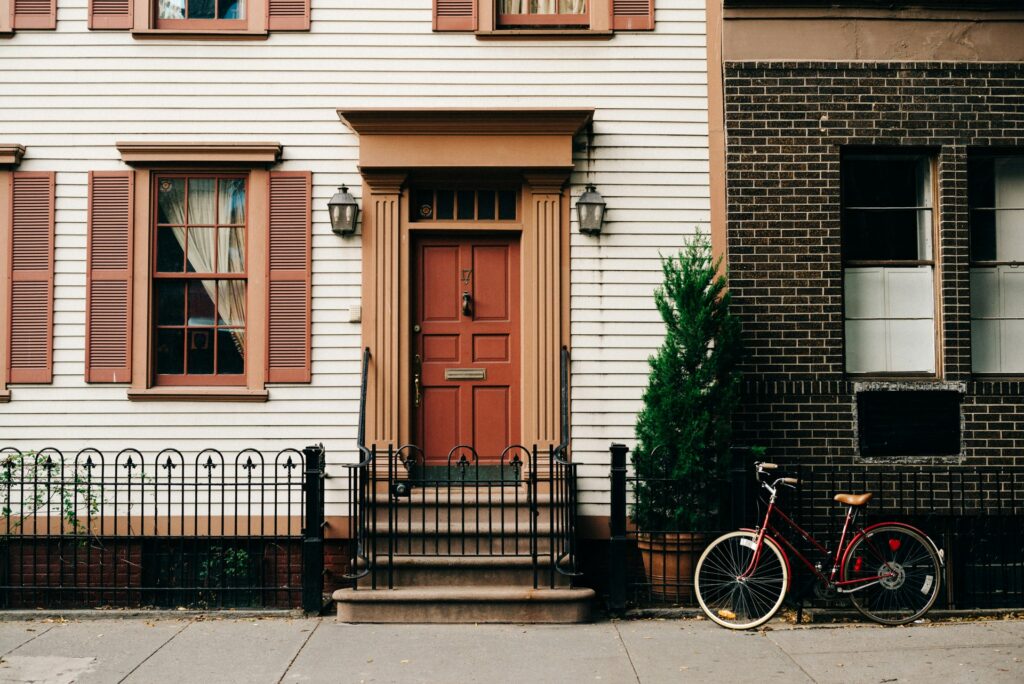 Front view of a house with a red door, brown shutters, and a small staircase. A black metal fence lines the sidewalk, and a red bicycle is parked next to a potted plant.