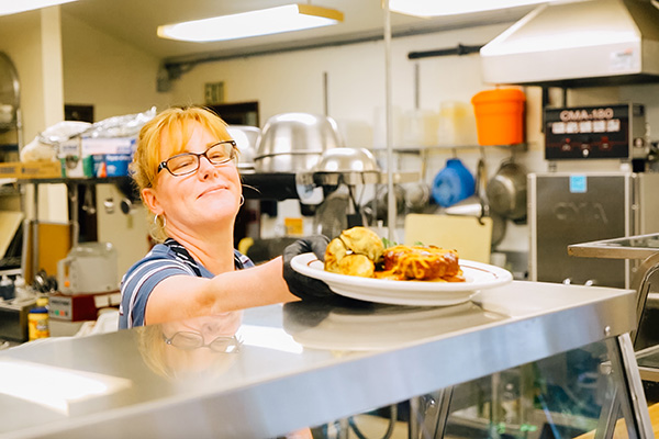 A person in a kitchen, wearing glasses and an apron, smiles while handing over a plate of food filled with various dishes. Kitchen equipment is visible in the background.