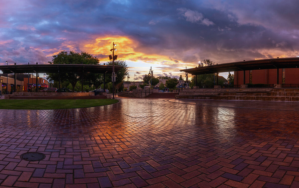 A wide plaza with wet brick pavement reflects a vibrant sunset. Covered seating areas and trees frame the scene. Cloudy sky with orange and purple hues overhead.