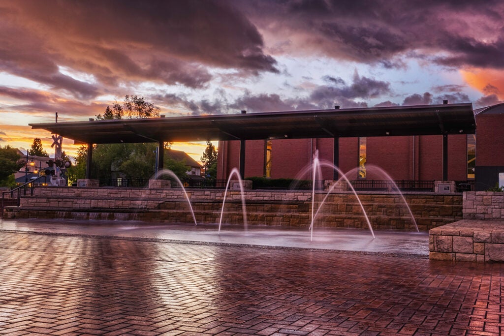 A brick courtyard features multiple arched water fountains. A covered structure and brick buildings are in the background, set against a dramatic sky during sunset.