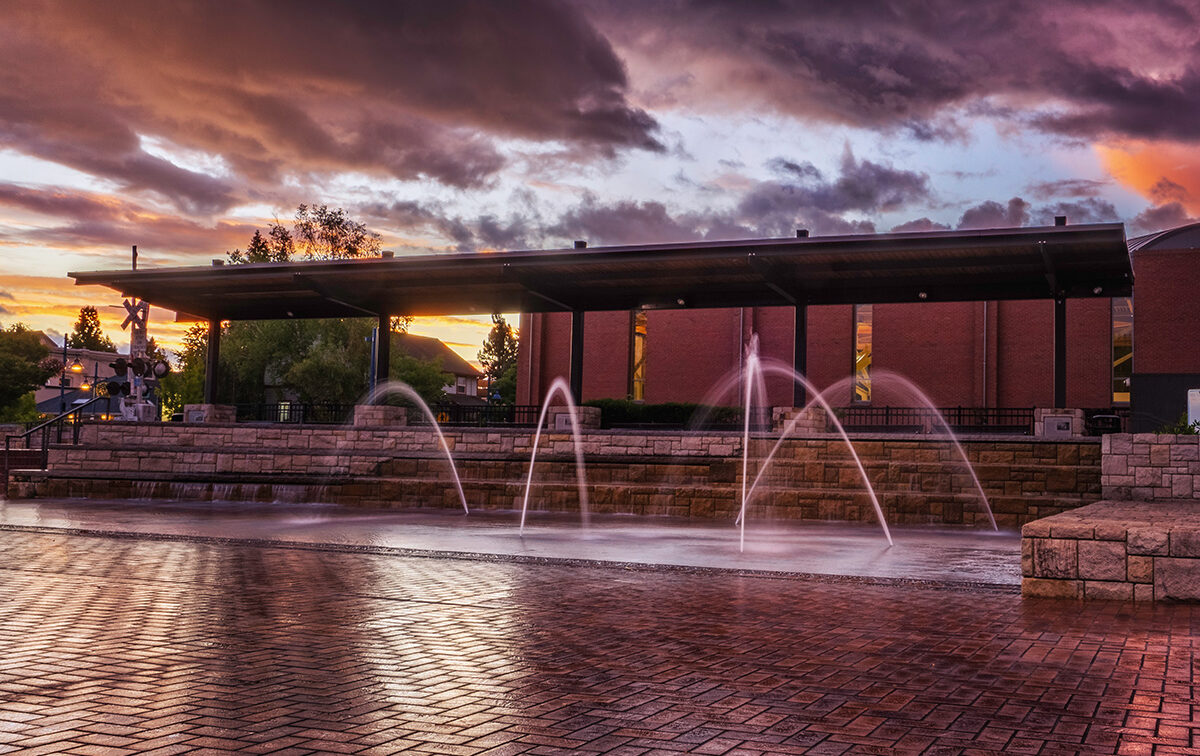 A brick courtyard features multiple arched water fountains. A covered structure and brick buildings are in the background, set against a dramatic sky during sunset.
