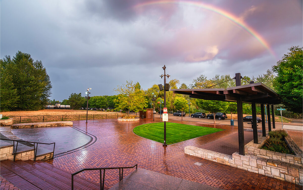 Brick-paved plaza with a grassy area, sheltered seating, and parked cars. Overcast sky with a rainbow. Trees and street in the background.