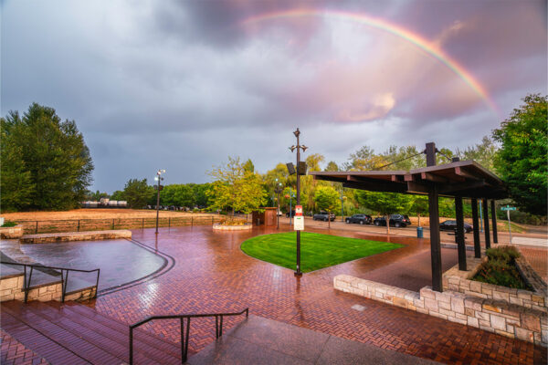 Brick-paved plaza with a grassy area, sheltered seating, and parked cars. Overcast sky with a rainbow. Trees and street in the background.