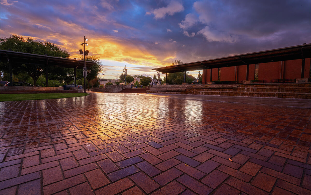 A wet brick plaza reflects a dramatic sunset sky. Trees and a building are visible in the background.