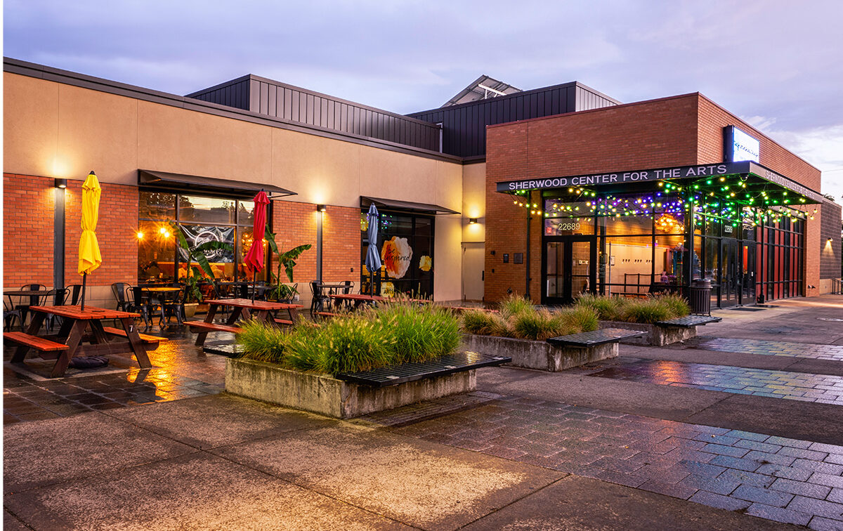 Outdoor view of Sherwood Center for the Arts entrance at dusk, with colorful string lights, potted plants, and seating area under umbrellas on a wet pavement.