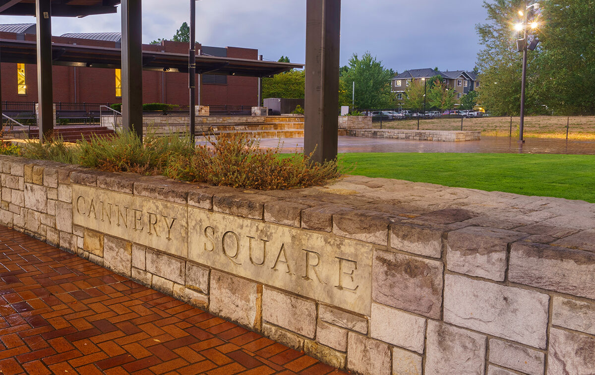 Brick and stone wall with the engraving "Cannery Square" in a landscaped outdoor area, featuring a modern building and trees in the background.