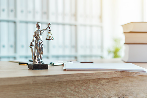 A small Lady Justice statue on a wooden desk with documents and stacked books in the background.