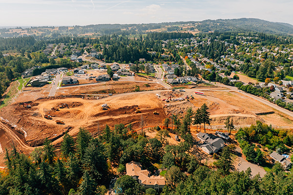 Aerial view of a cleared construction site surrounded by trees, with residential areas and distant hills visible in the background.