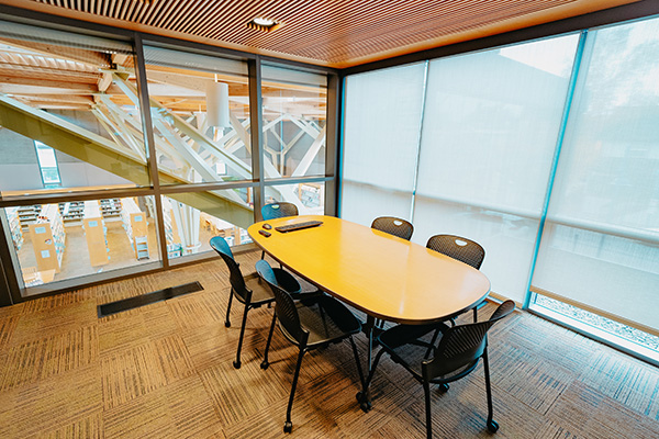 Meeting room with a wooden oval table and six black chairs, overlooking a library area through large windows. Blinds partially cover the windows.