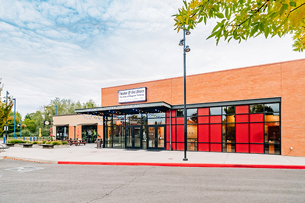 A modern library exterior with a brick facade, large windows, and a metal awning. The surroundings include trees and an outdoor seating area.