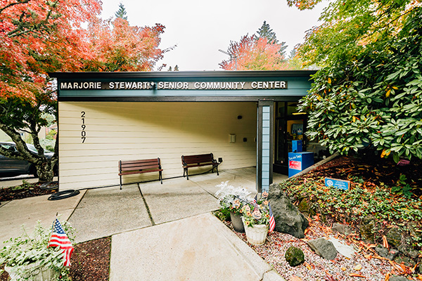 Exterior of Marjorie Stewart Senior Community Center with benches and American flags displayed, surrounded by autumn trees.