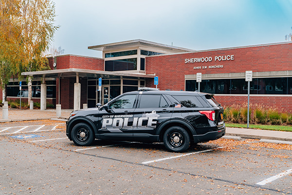 A police SUV is parked in front of the Sherwood Police Department building, surrounded by autumn leaves on the pavement.