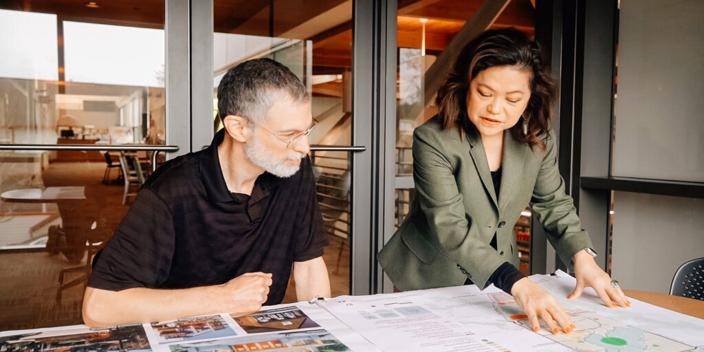 A man sitting and a woman standing, both reviewing architectural plans on a table in a modern office setting.