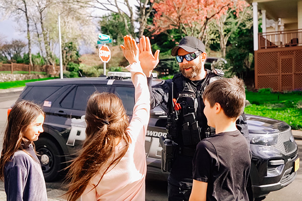 Police officer in uniform high-fives a child on a street near a police SUV, with two other children watching.