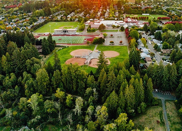 Aerial view of a town at sunset, featuring a sports complex with fields, trees, and residential areas. The sun is setting on the horizon, casting a warm glow over the landscape.