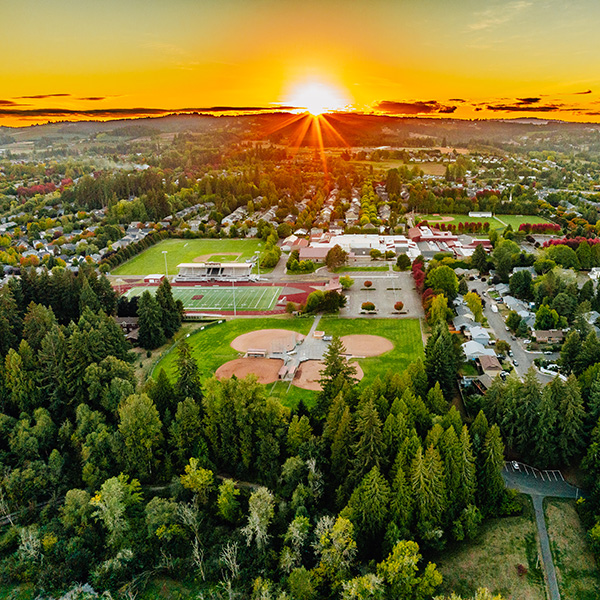 Aerial view of a town at sunset, featuring a sports complex with fields, trees, and residential areas. The sun is setting on the horizon, casting a warm glow over the landscape.