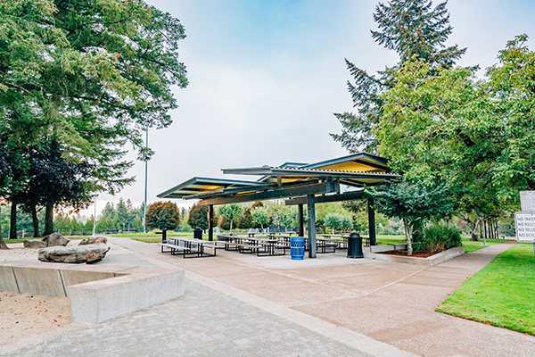 A covered outdoor pavilion with picnic tables and trash cans, surrounded by trees and a walkway in a park setting.