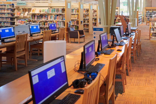 Rows of computers on wooden desks in a library, surrounded by bookshelves with various books; chairs are neatly arranged throughout the space.