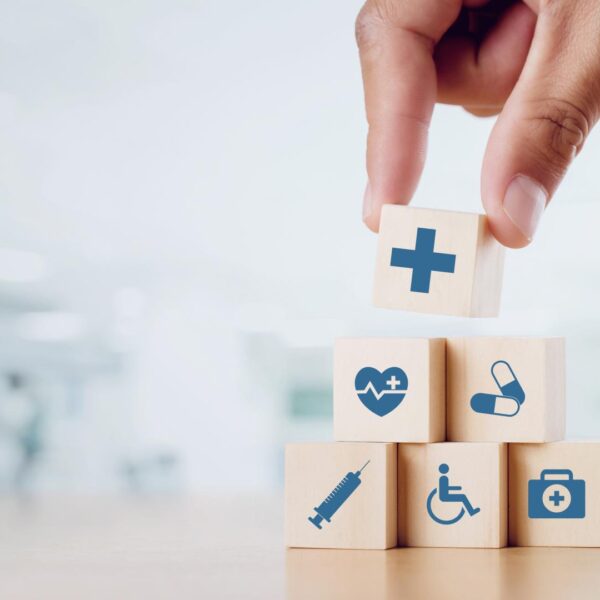 A hand stacks wooden blocks with medical icons, including a cross, heart, pills, syringe, wheelchair, and first aid kit, against a blurred healthcare setting background.