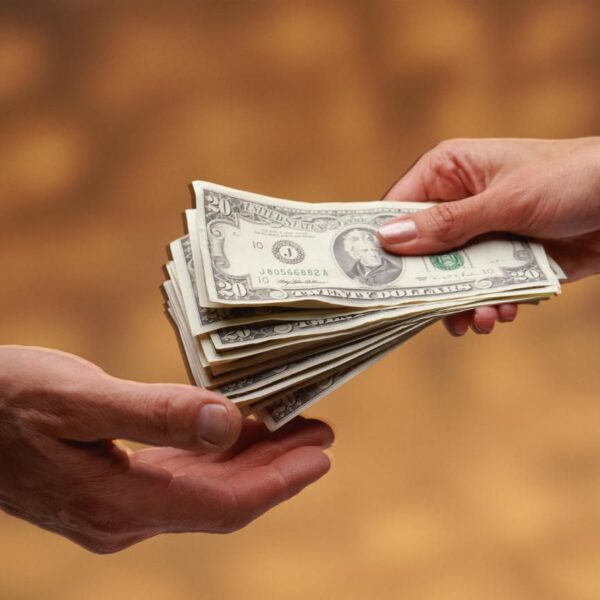 One person hands a stack of U.S. dollar bills to another person against a blurred brown background.