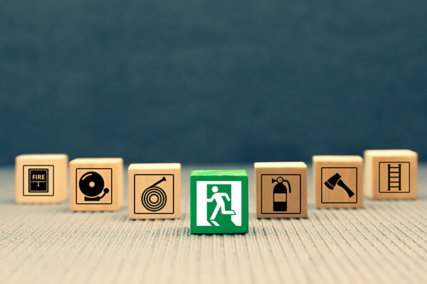 Seven wooden blocks with emergency symbols, including a fire alarm, hose, exit, fire extinguisher, axe, and ladder on a textured surface; the exit block is green, others are beige.
