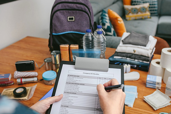 Person checking a preparedness checklist on a clipboard; items like water bottles, canned food, and first aid supplies are visible on the table.
