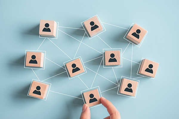 Wooden blocks with icons of people connected by lines on a blue background, representing a network or organizational structure. A hand reaches towards the blocks.