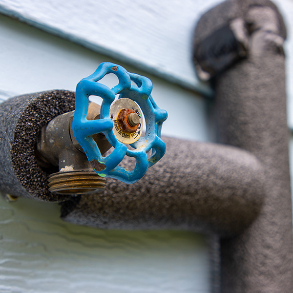 Close-up of an outdoor faucet with a blue handle, attached to a wall. The pipe is insulated with dark foam tubing.