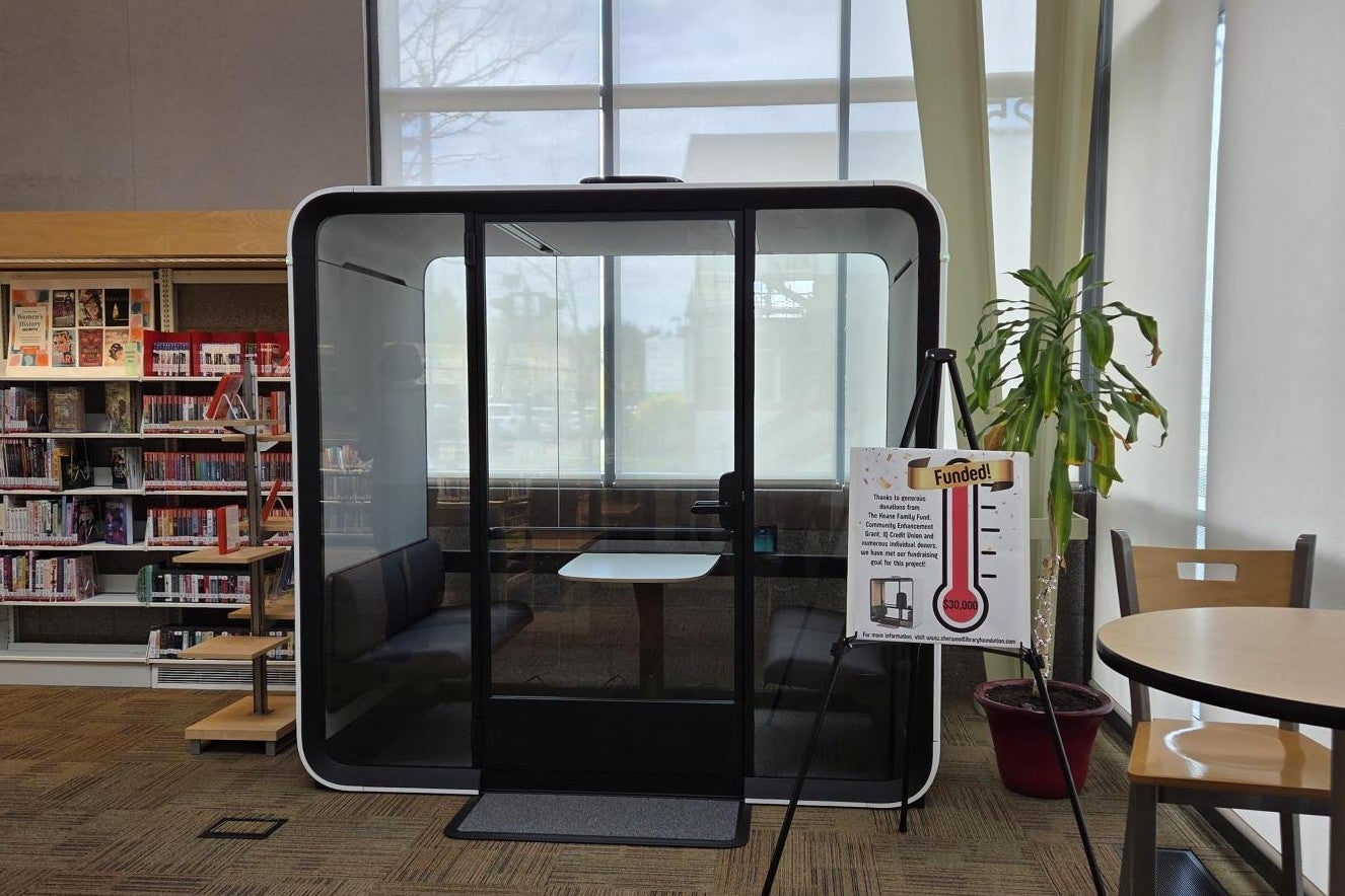 A glass-enclosed study pod sits in a library next to a thermometer fundraising sign, bookshelves, a potted plant, and a table with chairs.