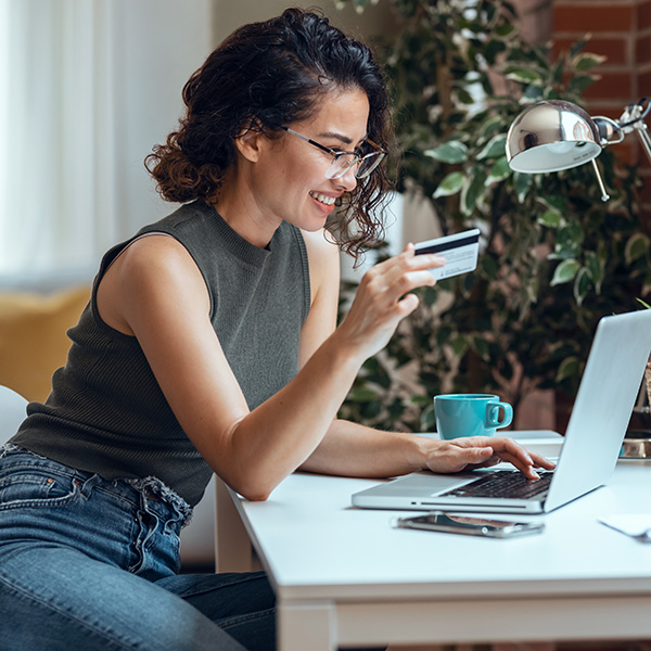 A woman with glasses holds a credit card and smiles while using a laptop at a desk with a smartphone, mug, and lamp nearby. A plant is in the background.