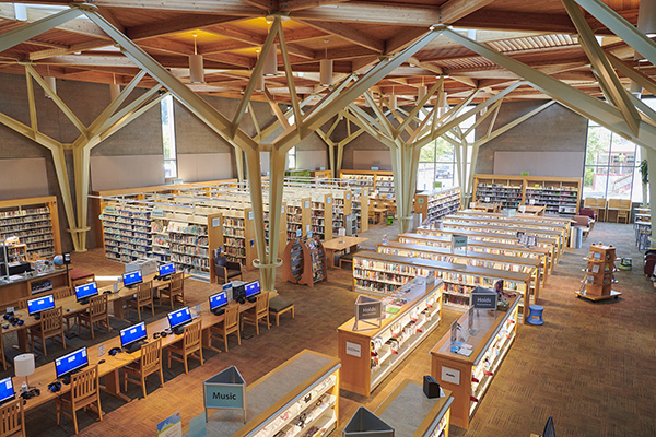 Spacious library interior with wooden beams, multiple bookshelves, computers on desks, and large windows allowing natural light.