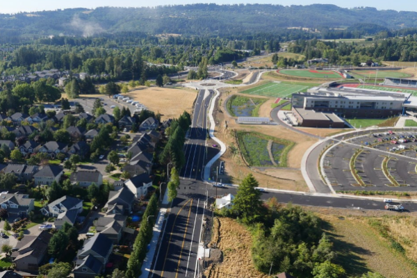 Aerial view of a suburban area with houses, a main road, a school building, athletic fields, and parking lots, surrounded by trees and hills.