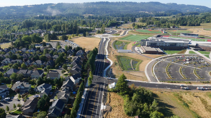 Aerial view of a suburban area with houses, a main road, a school building, athletic fields, and parking lots, surrounded by trees and hills.