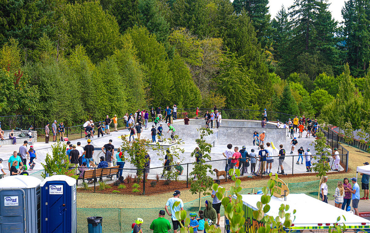 People gathering around and inside a skate park surrounded by trees, with portable toilets and tents visible nearby.