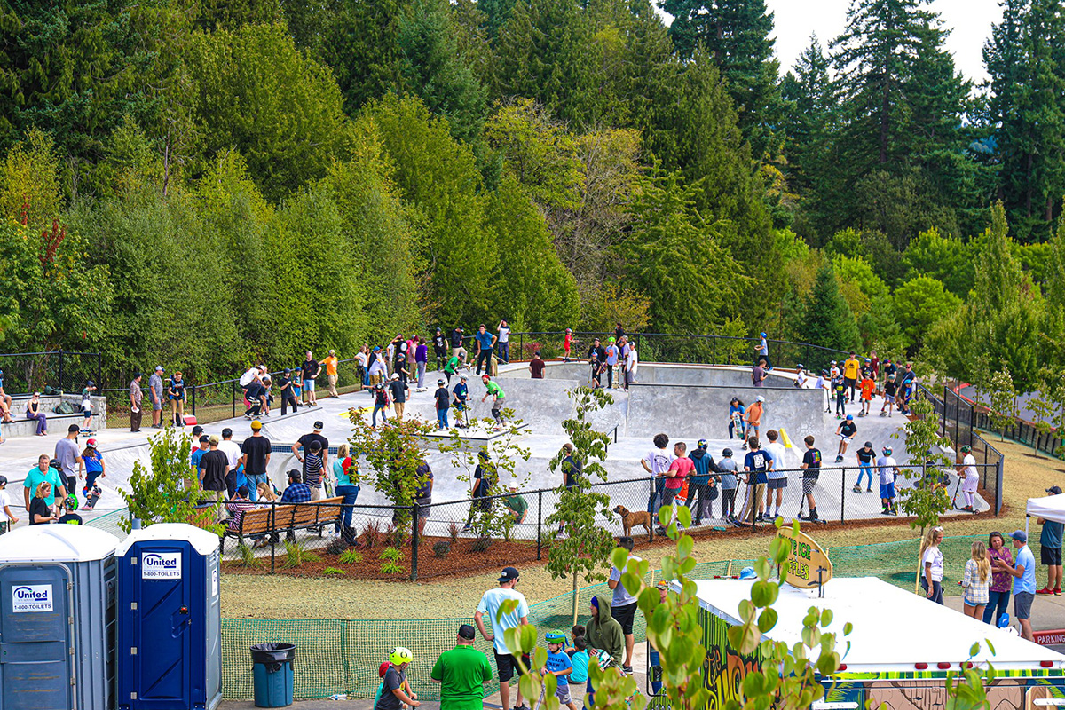 People gathering around and inside a skate park surrounded by trees, with portable toilets and tents visible nearby.