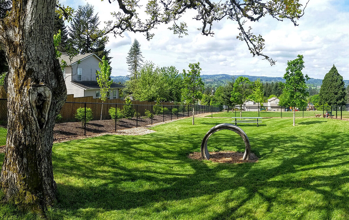A park landscape with a grassy field, trees, a wooden play tunnel, and houses beyond a fenced area under a partly cloudy sky.