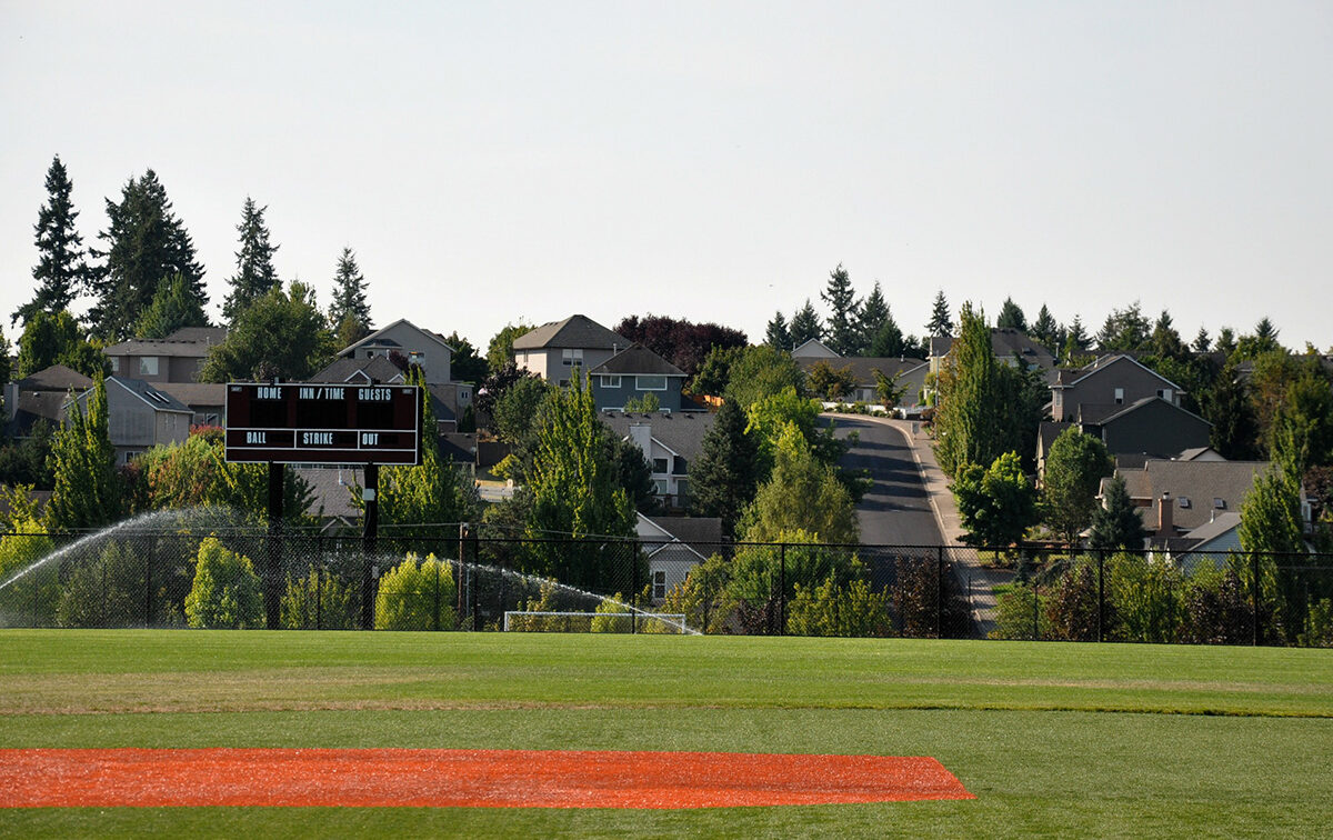 A baseball field with green grass and a red dirt area. A scoreboard and sprinklers are visible, with houses and trees lining a sloping street in the background.