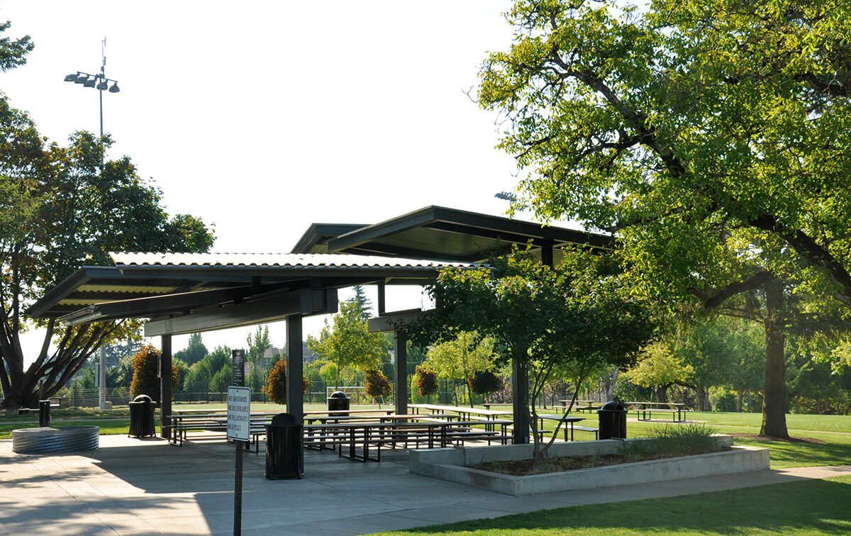 A park pavilion with metal roofing and picnic tables underneath, surrounded by green grass and trees in a sunlit outdoor setting.