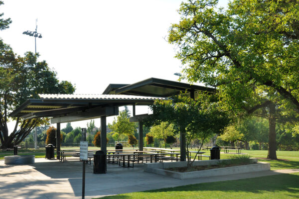 A park pavilion with metal roofing and picnic tables underneath, surrounded by green grass and trees in a sunlit outdoor setting.