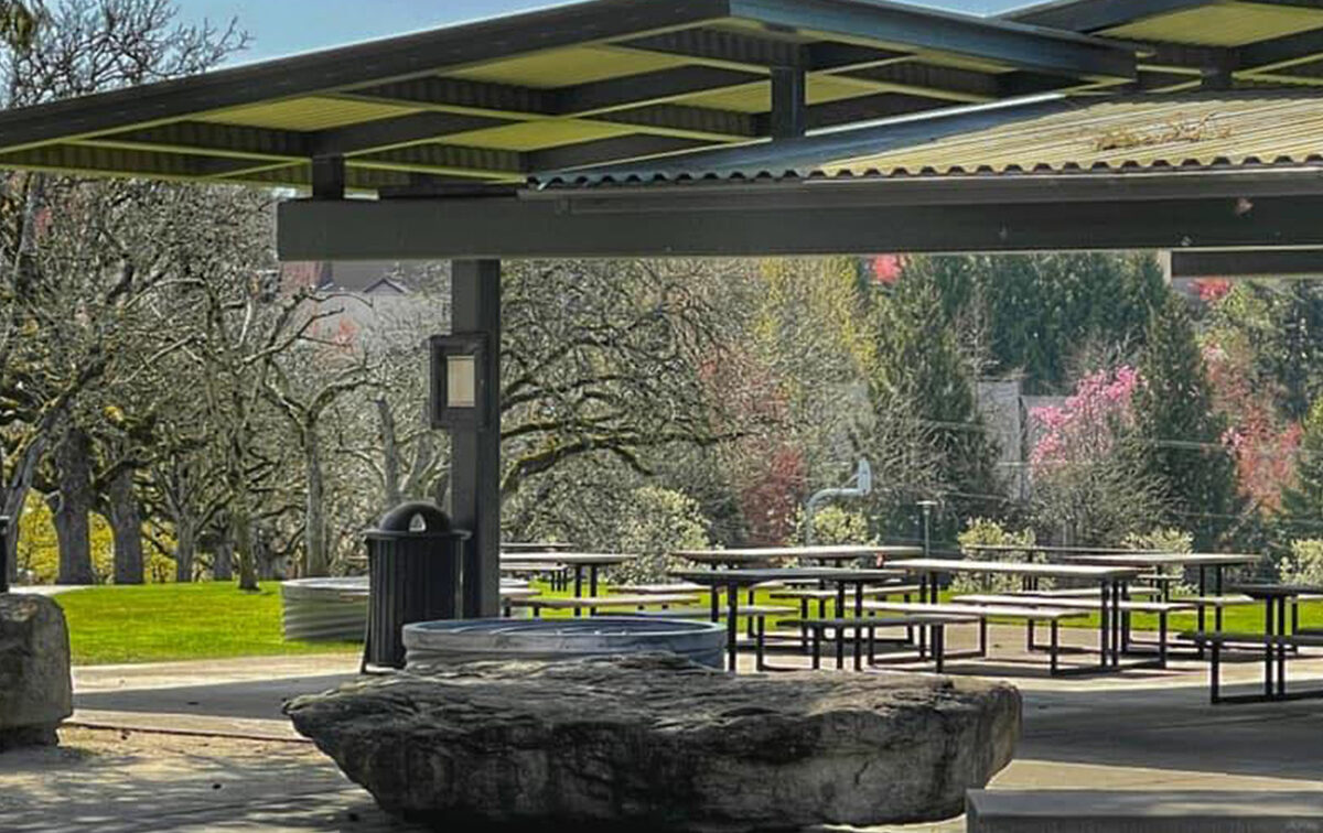 Outdoor pavilion with metal roof, picnic tables, and a rock bench. Trees and greenery are visible in the background on a clear day.