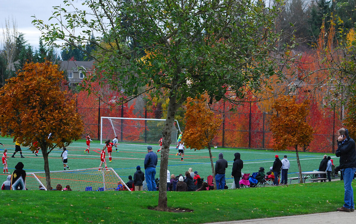 People watch a youth soccer game on a field surrounded by autumn trees, with a man taking photos on the sideline.