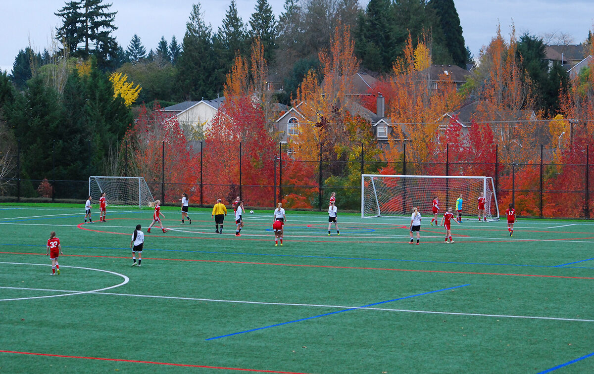 Kids playing soccer on a field surrounded by trees with autumn foliage, with a referee in yellow observing the game.