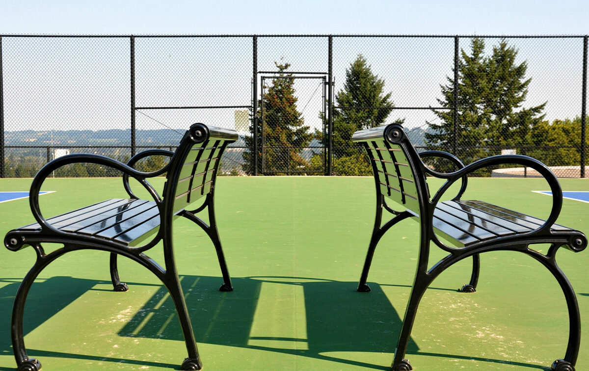 Two metal benches facing each other on a green outdoor court with a chain-link fence and trees in the background.