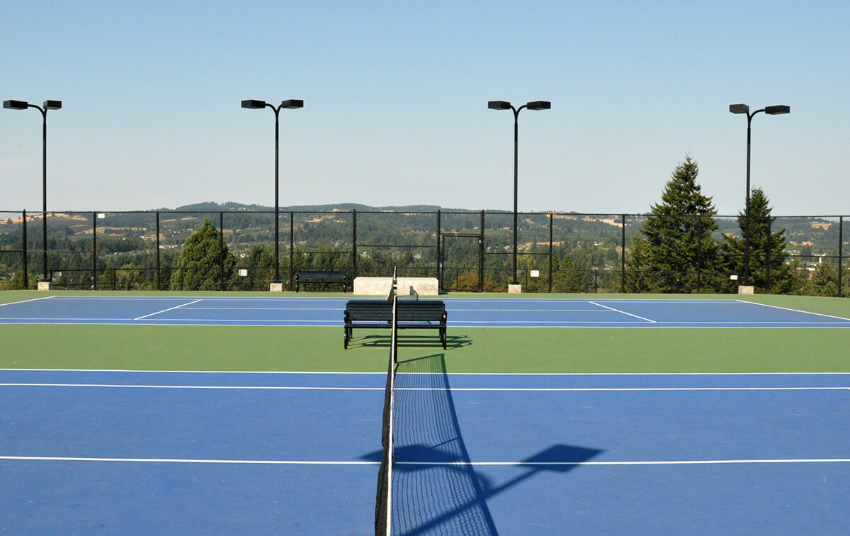 Outdoor tennis court with blue and green surface, surrounded by a black fence and trees in the background, under a clear sky. A bench sits in the center.