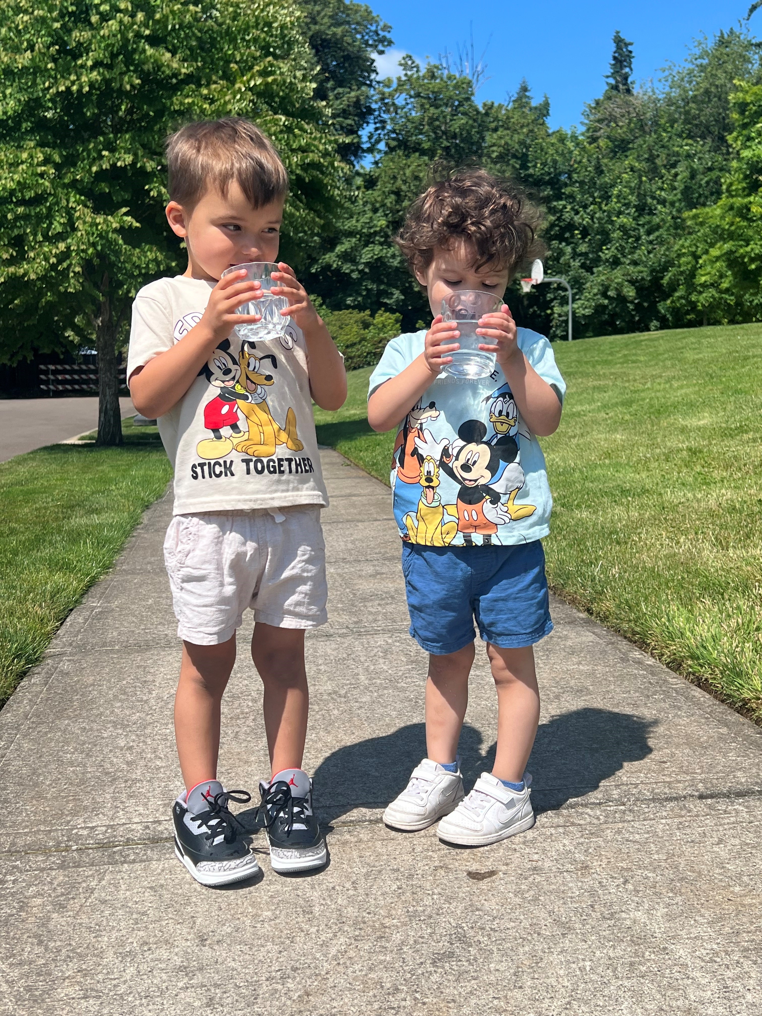 Two young children stand on a sidewalk outside, drinking from clear plastic cups. Both wear Mickey Mouse shirts and shorts, with grass and trees in the background.