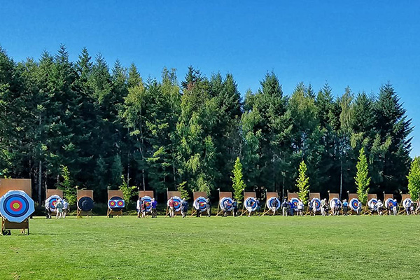 A row of archery targets set up on a grassy field with a backdrop of dense trees under a clear blue sky.